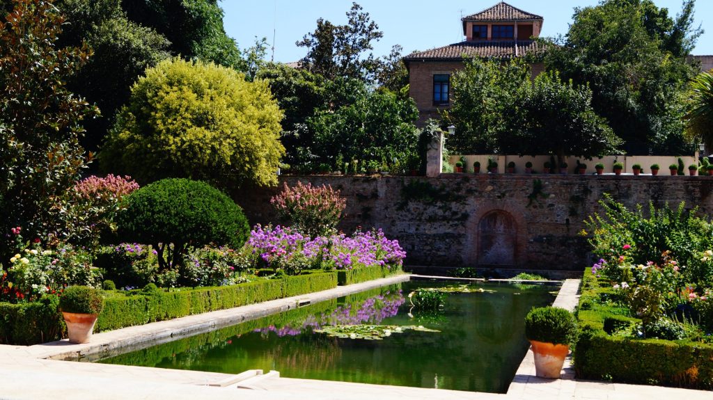 Lush garden with a rectangular pond, surrounded by greenery and flowers, with a building in the background.