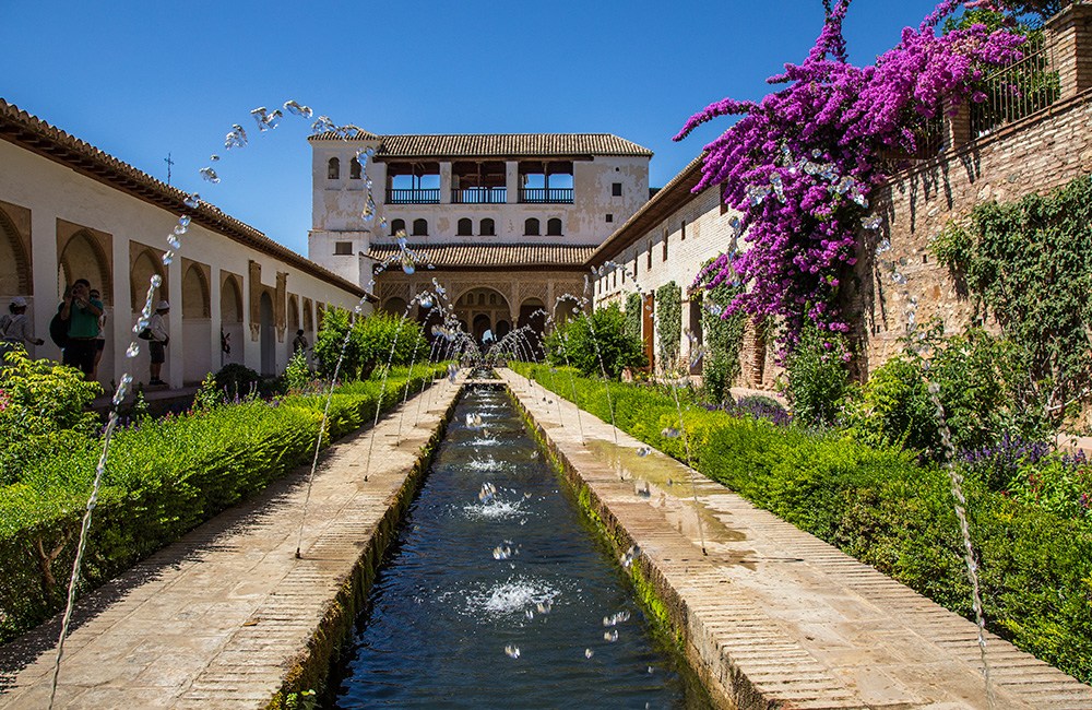 Garden with fountains, flowers, and historic building under blue sky.