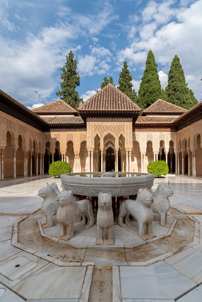 Courtyard with lion fountain, arches, and trees, under a blue sky with clouds.
