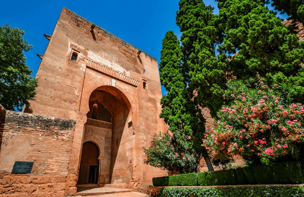 Historic stone archway with trees and flowering bushes under a clear blue sky.