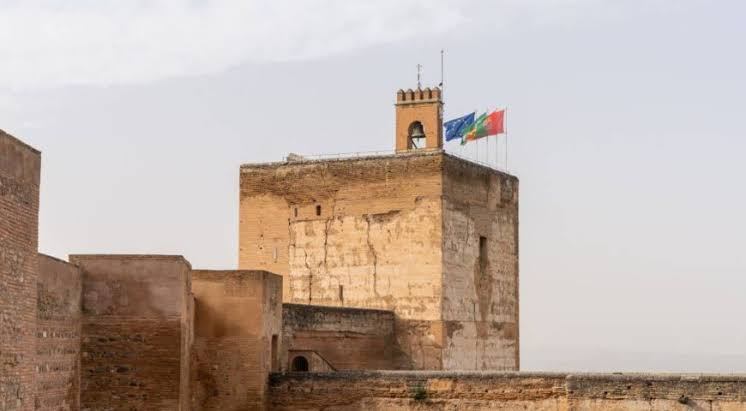 Historic stone tower with three flags and a cloudy sky background.