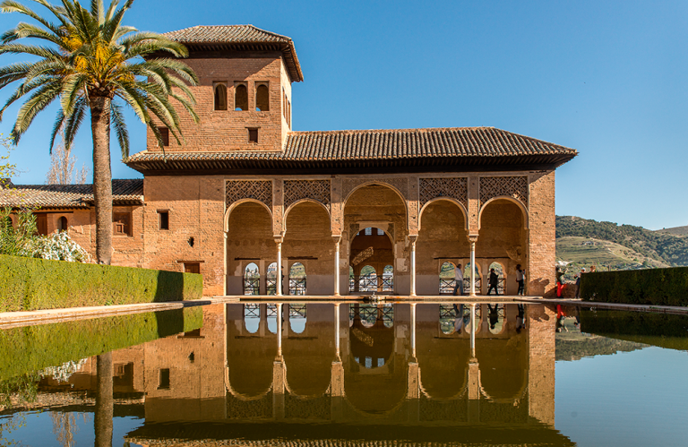 Historic building with arches reflected in a calm pond under a clear blue sky.