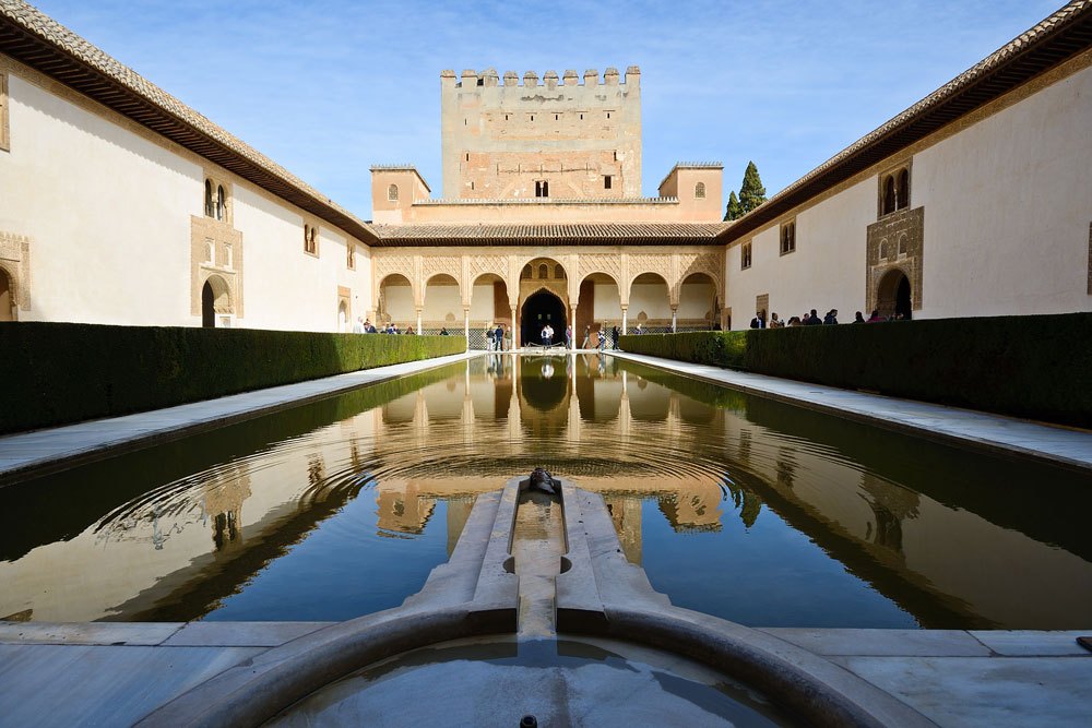 Courtyard with reflective pond and arched walkway under blue sky.